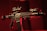 A black rifle with various attachments, including a scope and other tactical gear, is set against a dark, red-tinged background. The lighting emphasizes the structure of the weapon, casting shadows and highlighting its details.