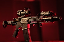 A black rifle with various attachments, including a scope and other tactical gear, is set against a dark, red-tinged background. The lighting emphasizes the structure of the weapon, casting shadows and highlighting its details.