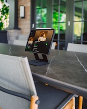 A tablet is placed on a stand on a large gray marble table. The screen displays a music app or playlist. The background features a patio with glass doors and green foliage visible through the windows.