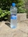 A plastic bottle of mineral water labeled 'Vatra,' with a blue cap, is standing on a light stone ground. The background features lush green leaves and a rough concrete block.