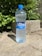 A plastic bottle of mineral water labeled 'Vatra,' with a blue cap, is standing on a light stone ground. The background features lush green leaves and a rough concrete block.