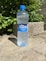 A plastic bottle of mineral water labeled 'Vatra,' with a blue cap, is standing on a light stone ground. The background features lush green leaves and a rough concrete block.