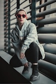 A fashionable individual crouches on a windowsill wearing a denim jacket, striped sleeves, and black pants. They have a scarf tied on their head and sport vibrant red sunglasses. The background features industrial-style window blinds.
