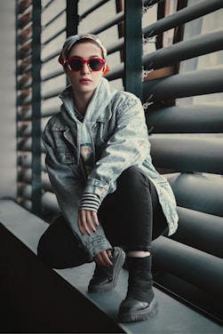 A fashionable individual crouches on a windowsill wearing a denim jacket, striped sleeves, and black pants. They have a scarf tied on their head and sport vibrant red sunglasses. The background features industrial-style window blinds.