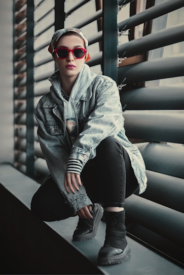 A fashionable individual crouches on a windowsill wearing a denim jacket, striped sleeves, and black pants. They have a scarf tied on their head and sport vibrant red sunglasses. The background features industrial-style window blinds.