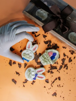 A gloved hand holds a chocolate with a bunny face design, surrounded by two more chocolates on a surface covered with chocolate shavings. The chocolates are designed with pastel-colored details resembling Easter themes, such as a basket with eggs. A box with a bow containing more chocolates is partially visible in the background.