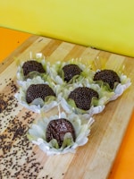 A wooden board displays several chocolate truffles in decorative paper wrappings resembling flowers. Chocolate sprinkles are scattered around, and the background features a bright yellow wall.