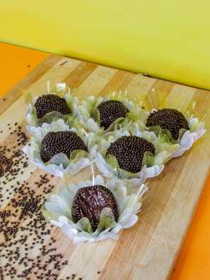 A wooden board displays several chocolate truffles in decorative paper wrappings resembling flowers. Chocolate sprinkles are scattered around, and the background features a bright yellow wall.