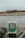 A makeshift trash receptacle labeled 'BUTT HUT' stands on a rocky shore. The container is painted teal and white, topped with a simple, rustic wooden roof. The background is overcast, with a sandy beach and distant trees.