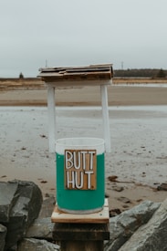 A makeshift trash receptacle labeled 'BUTT HUT' stands on a rocky shore. The container is painted teal and white, topped with a simple, rustic wooden roof. The background is overcast, with a sandy beach and distant trees.