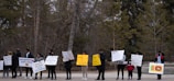 A group of people standing outdoors holding various protest signs. The background consists of trees without leaves, indicating a winter or early spring setting. The signs include messages about supporting Sri Lanka, political leadership, and other slogans.