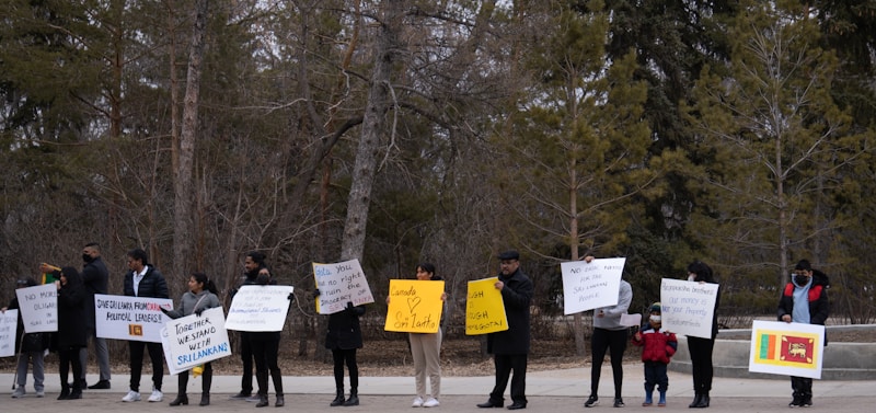 A group of people standing outdoors holding various protest signs. The background consists of trees without leaves, indicating a winter or early spring setting. The signs include messages about supporting Sri Lanka, political leadership, and other slogans.