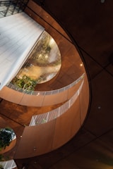 An architectural interior featuring curved walkways with white railings, surrounded by warm-toned walls. Green plants are visible in the structure, adding a natural element to the modern design. Large, circular openings create an airy and open feeling, while reflections play on the surfaces.