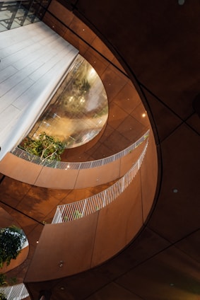 An architectural interior featuring curved walkways with white railings, surrounded by warm-toned walls. Green plants are visible in the structure, adding a natural element to the modern design. Large, circular openings create an airy and open feeling, while reflections play on the surfaces.