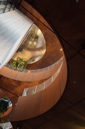 An architectural interior featuring curved walkways with white railings, surrounded by warm-toned walls. Green plants are visible in the structure, adding a natural element to the modern design. Large, circular openings create an airy and open feeling, while reflections play on the surfaces.