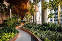 A lush green garden pathway with dense foliage of various plants and trees, surrounded by modern, sleek architectural columns. The pathway curves gently and is well-lit with warm lighting, creating an inviting and serene atmosphere.