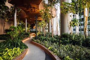 A lush green garden pathway with dense foliage of various plants and trees, surrounded by modern, sleek architectural columns. The pathway curves gently and is well-lit with warm lighting, creating an inviting and serene atmosphere.
