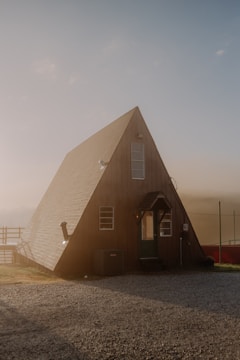 An A-frame cabin with wood paneling and a steeply sloped roof stands on a gravel lot. The structure features multiple windows, including a large vertical window and a small porch at the front entrance. The setting appears to be rural with a slightly misty atmosphere in the background and a soft, diffused light illuminating the scene.