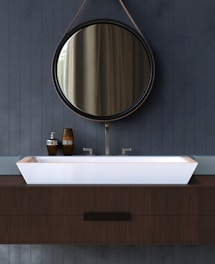 A modern bathroom vanity with a circular mirror hanging above a rectangular white sink. The mirror is framed with a thin, dark rim and suspended by a leather strap. The sink rests on a wooden countertop, complemented by metallic faucets. On the left side of the sink, there are brown and silver soap dispensers.