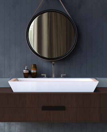 A modern bathroom vanity with a circular mirror hanging above a rectangular white sink. The mirror is framed with a thin, dark rim and suspended by a leather strap. The sink rests on a wooden countertop, complemented by metallic faucets. On the left side of the sink, there are brown and silver soap dispensers.
