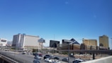 A cityscape featuring several large hotels and casinos, including buildings with recognizable names. These structures have bright exteriors and are set against a clear blue sky. Below, a freeway with moving traffic is visible, showing numerous cars.