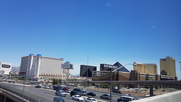 A cityscape featuring several large hotels and casinos, including buildings with recognizable names. These structures have bright exteriors and are set against a clear blue sky. Below, a freeway with moving traffic is visible, showing numerous cars.