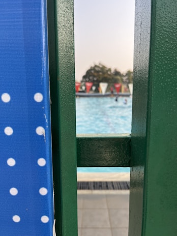 A close-up view through a green metal fence showing a swimming pool in the background. There are several people in the pool and colorful triangular flags are hanging on a line across the pool. To the left, part of a blue surface with white polka dots is visible.