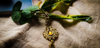 An ornate, gold-colored jewelry piece with intricate designs, featuring a central gemstone, is elegantly placed on a textured fabric surface. Green leaves and a partially visible book provide a natural and serene backdrop.