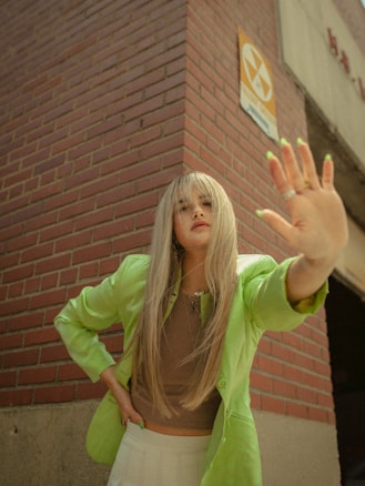 A person with long blonde hair is wearing a bright green jacket and a brown top, standing with one arm extended towards the camera. They appear to be against a brick wall with an aged radiation sign above.