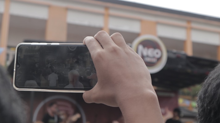 A person is holding a smartphone horizontally and recording a scene that appears to be a live outdoor performance or event. There is a partially visible stage with a band or performers in the background. Above the stage is an out-of-focus sign with the text 'Neo.' The surroundings include parts of a building and other attendees.