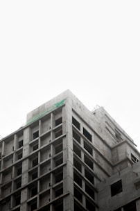 A partially constructed multi-story concrete building with an exposed framework. The structure is layered with visible grids of columns and beams, suggesting early stages of construction. The sky above appears overcast, creating a muted backdrop.