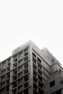 A partially constructed multi-story concrete building with an exposed framework. The structure is layered with visible grids of columns and beams, suggesting early stages of construction. The sky above appears overcast, creating a muted backdrop.