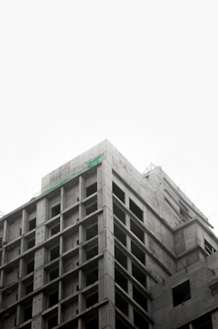 A partially constructed multi-story concrete building with an exposed framework. The structure is layered with visible grids of columns and beams, suggesting early stages of construction. The sky above appears overcast, creating a muted backdrop.
