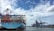 A large cargo ship loaded with multicolored shipping containers is docked at a busy port. Massive cranes with red and blue structures stand along the shoreline, used for loading and unloading containers. The sky is partly cloudy, and the water is calm, reflecting the cranes and ship.