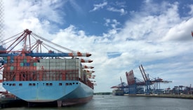 A large cargo ship loaded with multicolored shipping containers is docked at a busy port. Massive cranes with red and blue structures stand along the shoreline, used for loading and unloading containers. The sky is partly cloudy, and the water is calm, reflecting the cranes and ship.