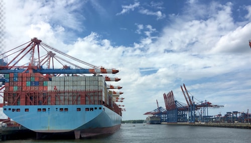 A large cargo ship loaded with multicolored shipping containers is docked at a busy port. Massive cranes with red and blue structures stand along the shoreline, used for loading and unloading containers. The sky is partly cloudy, and the water is calm, reflecting the cranes and ship.
