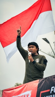 A person wearing a dark long-sleeved shirt is speaking into a microphone, raising their index finger into the air. They are standing in front of a large red and white flag flying overhead. The sky is overcast, and there appears to be a streetlight or pole in the background.