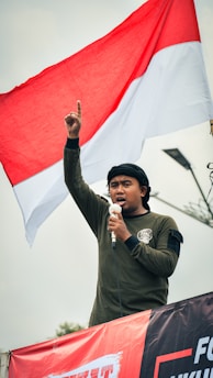 A person wearing a dark long-sleeved shirt is speaking into a microphone, raising their index finger into the air. They are standing in front of a large red and white flag flying overhead. The sky is overcast, and there appears to be a streetlight or pole in the background.