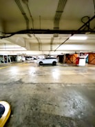 A car is parked in a well-lit parking garage with visible ceiling pipes and a concrete floor. The parking area is marked with yellow lines and a red sign indicating a parking level.