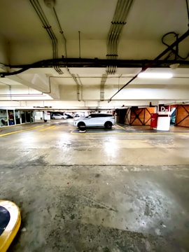 A car is parked in a well-lit parking garage with visible ceiling pipes and a concrete floor. The parking area is marked with yellow lines and a red sign indicating a parking level.