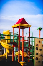 A vibrant playground structure featuring a spiral slide, climbing wall, and various play equipment. The bright colors are highlighted under a clear blue sky, with distinctive reds, yellows, and greens making up the play area.