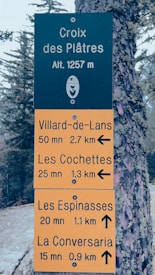 A signboard indicating directions and distances to various locations. The top section of the sign is black with white text and a floral emblem. The lower section is yellow with black text providing distances and estimated times to Villard-de-Lans, Les Cochettes, Les Espinasses, and La Conversaria. The background includes trees and a tree trunk with moss.