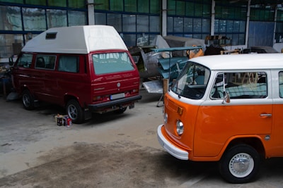 Two vintage Volkswagen vans are parked inside a workshop. The van in the foreground has a bright orange and white exterior, while the one behind is red with a white roof. The workshop is cluttered with various car parts and tools. The floor is concrete, and the walls are lined with industrial-style windows.