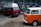 Two vintage Volkswagen vans are parked inside a workshop. The van in the foreground has a bright orange and white exterior, while the one behind is red with a white roof. The workshop is cluttered with various car parts and tools. The floor is concrete, and the walls are lined with industrial-style windows.