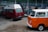 Two vintage Volkswagen vans are parked inside a workshop. The van in the foreground has a bright orange and white exterior, while the one behind is red with a white roof. The workshop is cluttered with various car parts and tools. The floor is concrete, and the walls are lined with industrial-style windows.