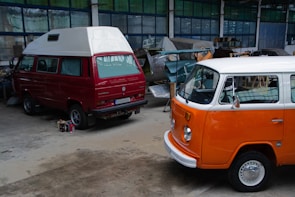 Two vintage Volkswagen vans are parked inside a workshop. The van in the foreground has a bright orange and white exterior, while the one behind is red with a white roof. The workshop is cluttered with various car parts and tools. The floor is concrete, and the walls are lined with industrial-style windows.