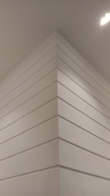A corner of a modern interior space with smooth white paneling extending vertically. The lighting casts soft shadows, giving a sense of depth. A bright ceiling light is visible, illuminating the area.
