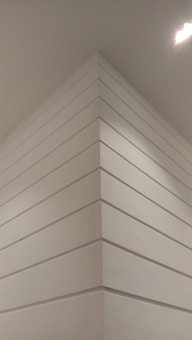 A corner of a modern interior space with smooth white paneling extending vertically. The lighting casts soft shadows, giving a sense of depth. A bright ceiling light is visible, illuminating the area.