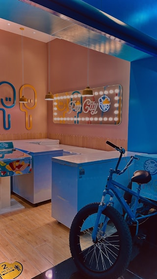 A shop interior with ice cream freezers and a decorative neon sign on the wall. The sign has the words 'Pop City' and features popsicle designs. A blue bicycle is parked next to the freezers. The lighting is warm, giving the space a cozy feel.