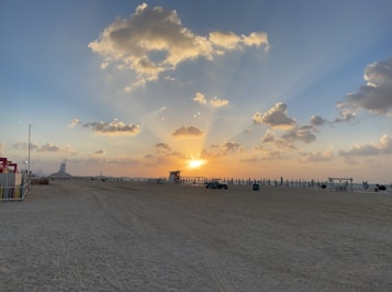 A serene beach scene with a prominent sunset creating dramatic rays of light through scattered clouds. The sandy beach is expansive, with few structures and a distant iconic building visible on the horizon. Beach umbrellas are neatly organized along the shoreline.